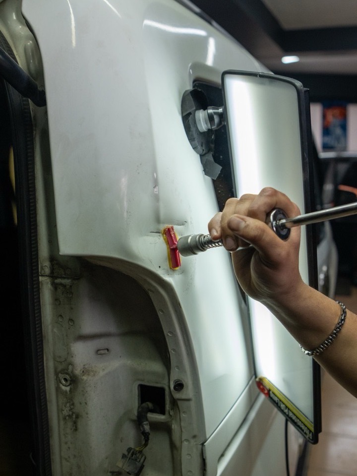 A technician using a paintless dent repair tool on a white van panel, checking the dent against a light board.