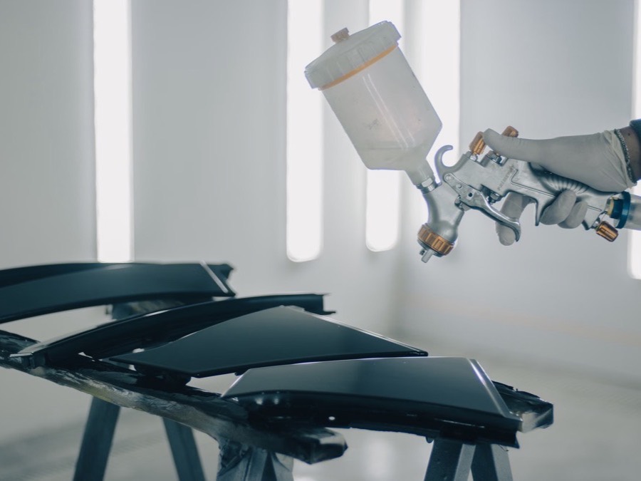 A spray gun applying black paint to car body panels inside a bright paint booth.